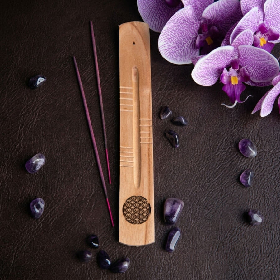 Handgefertigter Räucherstäbchenhalter in Schiffchenform aus Mangoholz mit Schnitzereien und Lasergravur der Blume des Lebens auf einem Hintergrund mit Ledertextur. Danaben liegen einige Räucherstäbchen, Amethyst Trommelsteine und Orchideenblüten als Dekoelemente.