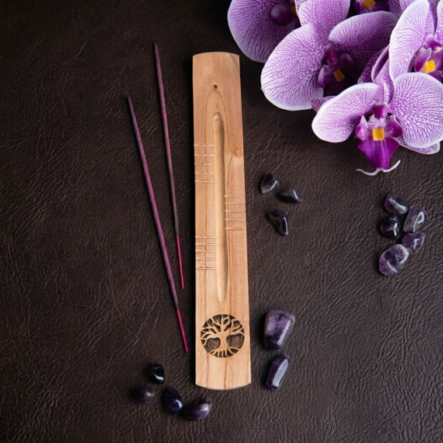 Handgefertigter Räucherstäbchenhalter in Schiffchenform aus Mangoholz mit Schnitzereien und Yggdrasil als Lasergravur auf einem Hintergrund mit Ledertextur. Danaben liegen einige Räucherstäbchen, Amethyst Trommelsteine und Orchideenblüten als Dekoelemente.