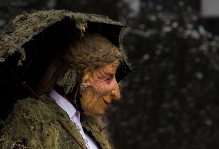 Dieses Bild zeigt eine handgefertigte, altehrwürdige Perchten- oder Hexenmaske mit faltiger Haut, spitzer Nase und einem moosbedeckten Hut, wie sie oft im alpinen Brauchtum während der Rauhnächte verwendet wird.
